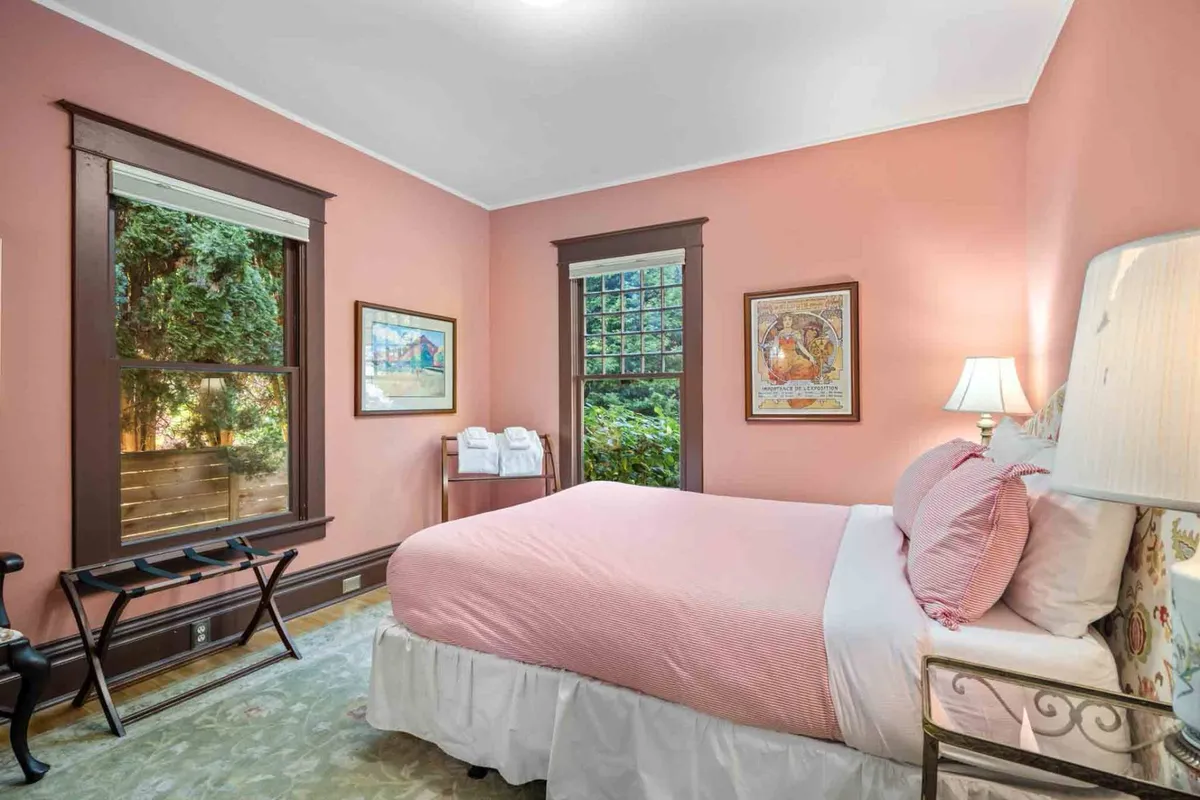A cozy bedroom featuring a plush bed dressed in light linens, a bedside lamp casting a gentle glow, a window allowing natural light to filter in, and tasteful artwork on the wall. The space is intimate and tranquil, designed for rest and relaxation.