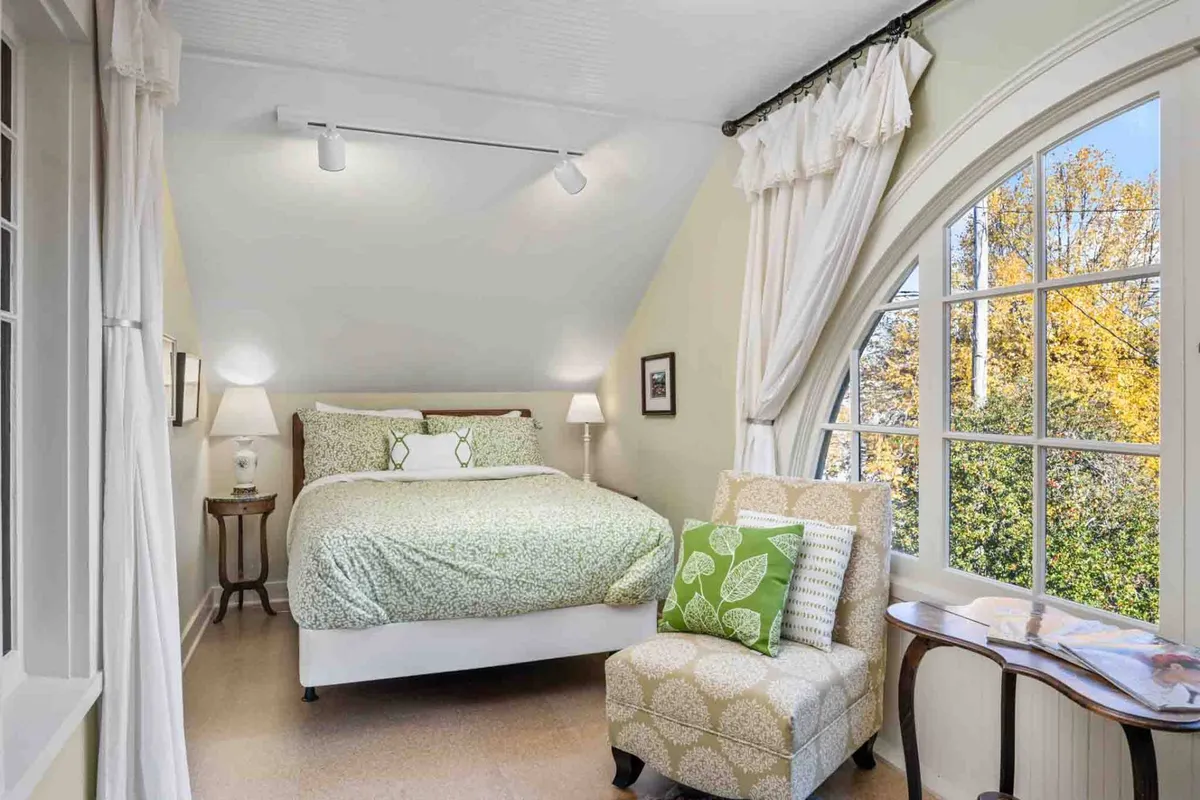 A tranquil bedroom that features a bed with green patterned bedding, a cozy armchair with a leafy design positioned by a large arched window, and soft filtered daylight streaming in. The room is inviting for both relaxation and reading.