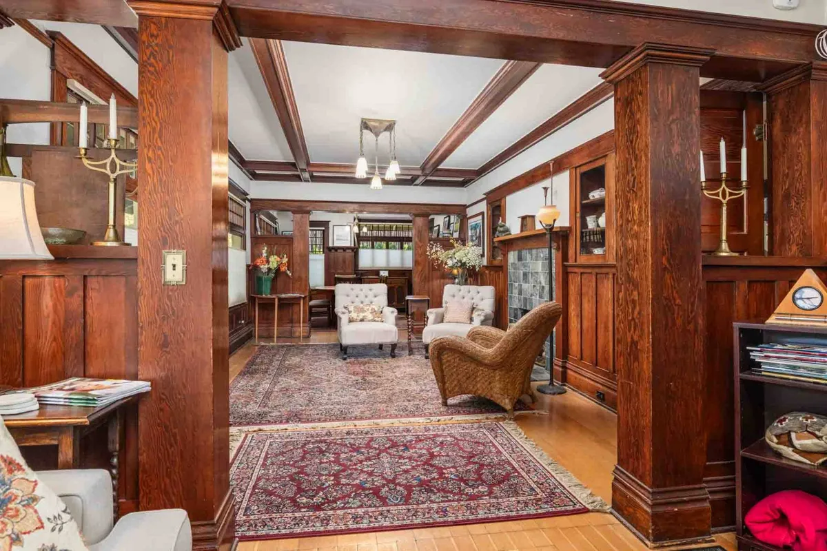 A warm, inviting living room with exposed wooden beams spanning the ceiling, a central fireplace flanked by comfortable seating, an area rug, and period accents. The arrangement encourages conversation, relaxation, and group gatherings.