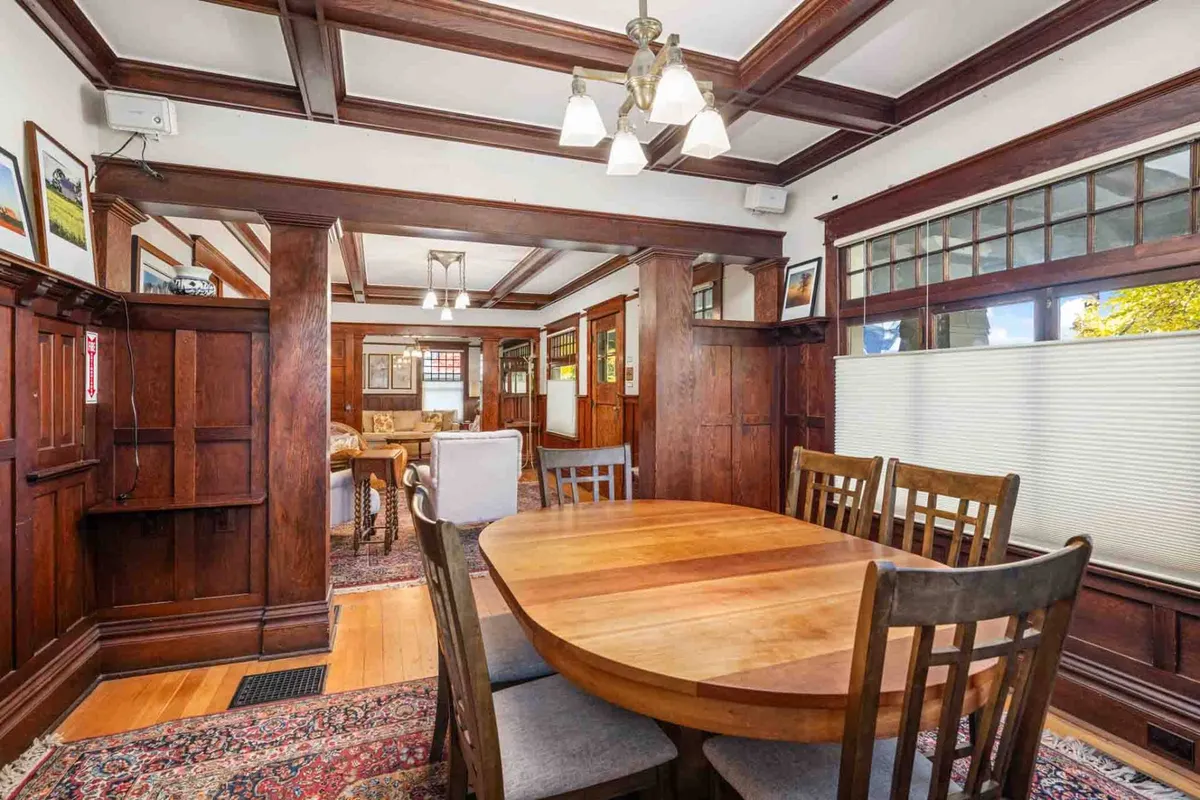 A warmly lit dining room featuring a large wooden table set for eight, with a beamed ceiling, chandelier, wood paneling, and windows that invite natural light into the space. The setting exudes a cozy, welcoming atmosphere perfect for family dinners and celebratory gatherings.