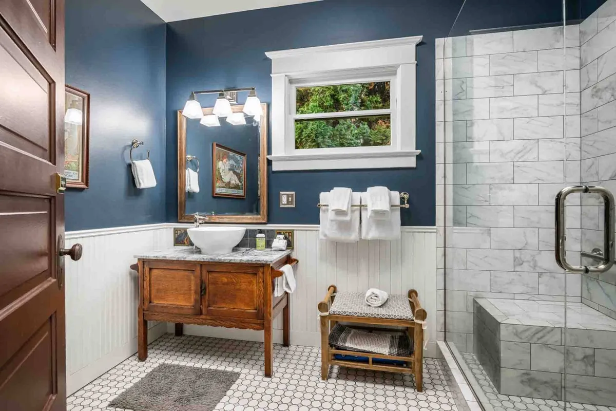 A stylish, updated bathroom with a vintage wooden vanity, marble shower, crisp white towels, and framed artwork, combining modern comfort with historic charm. The space is bright and inviting, ideal for pampering and self-care.