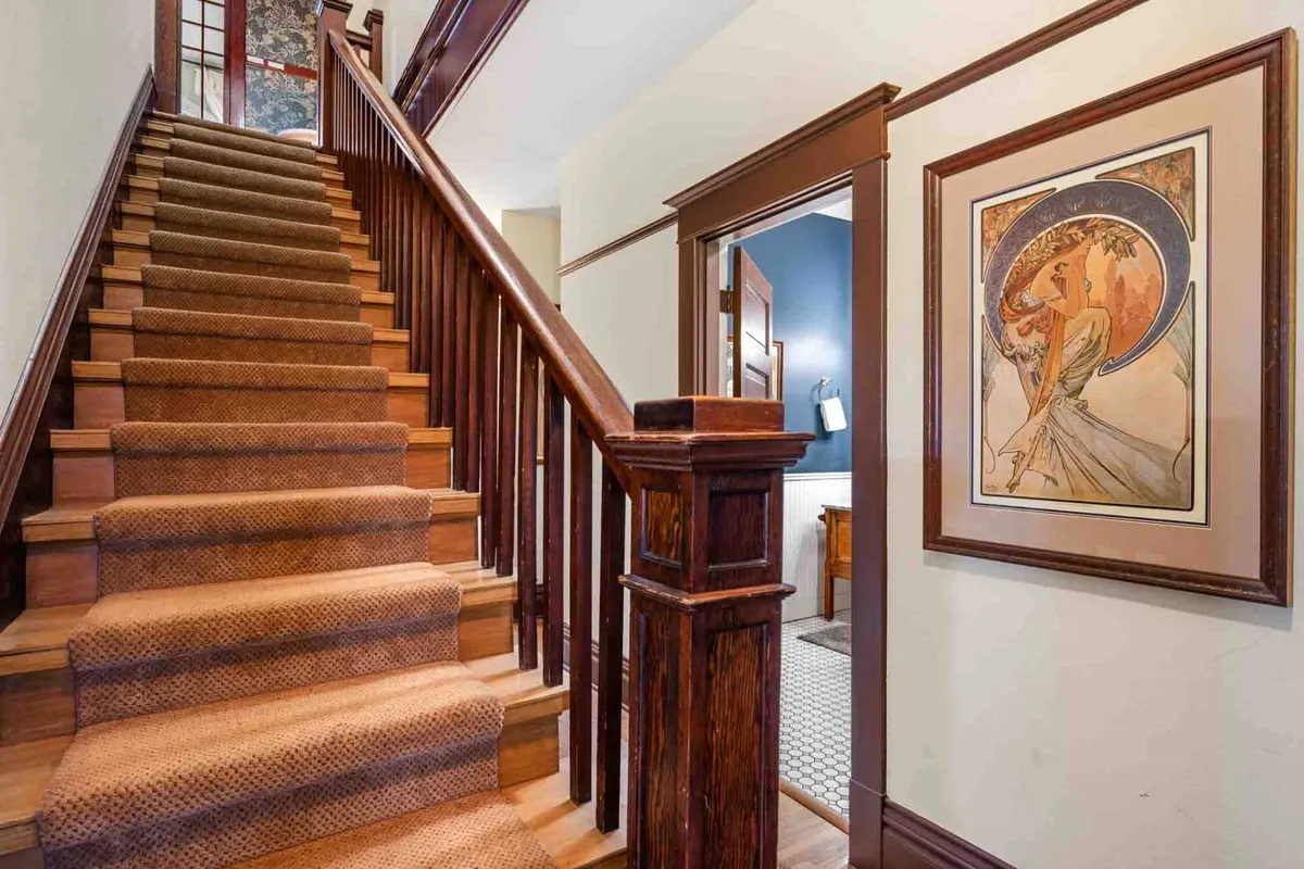 The home's staircase area is adorned with artwork and features a welcoming wooden railing, creating a warm transition space ideal for guests moving between rooms or gathering in small groups. The inviting hallway suggests discovery and togetherness as guests navigate shared spaces.