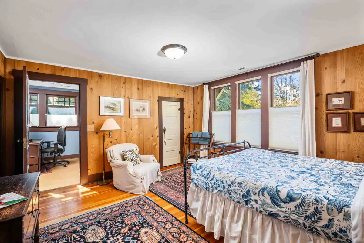 A serene bedroom with floral bedding, a comfortable chair positioned by the window, and artwork on the walls. Natural light pours in through the windows, illuminating a space that is both restful and conducive to reading, working, or simply enjoying the view.