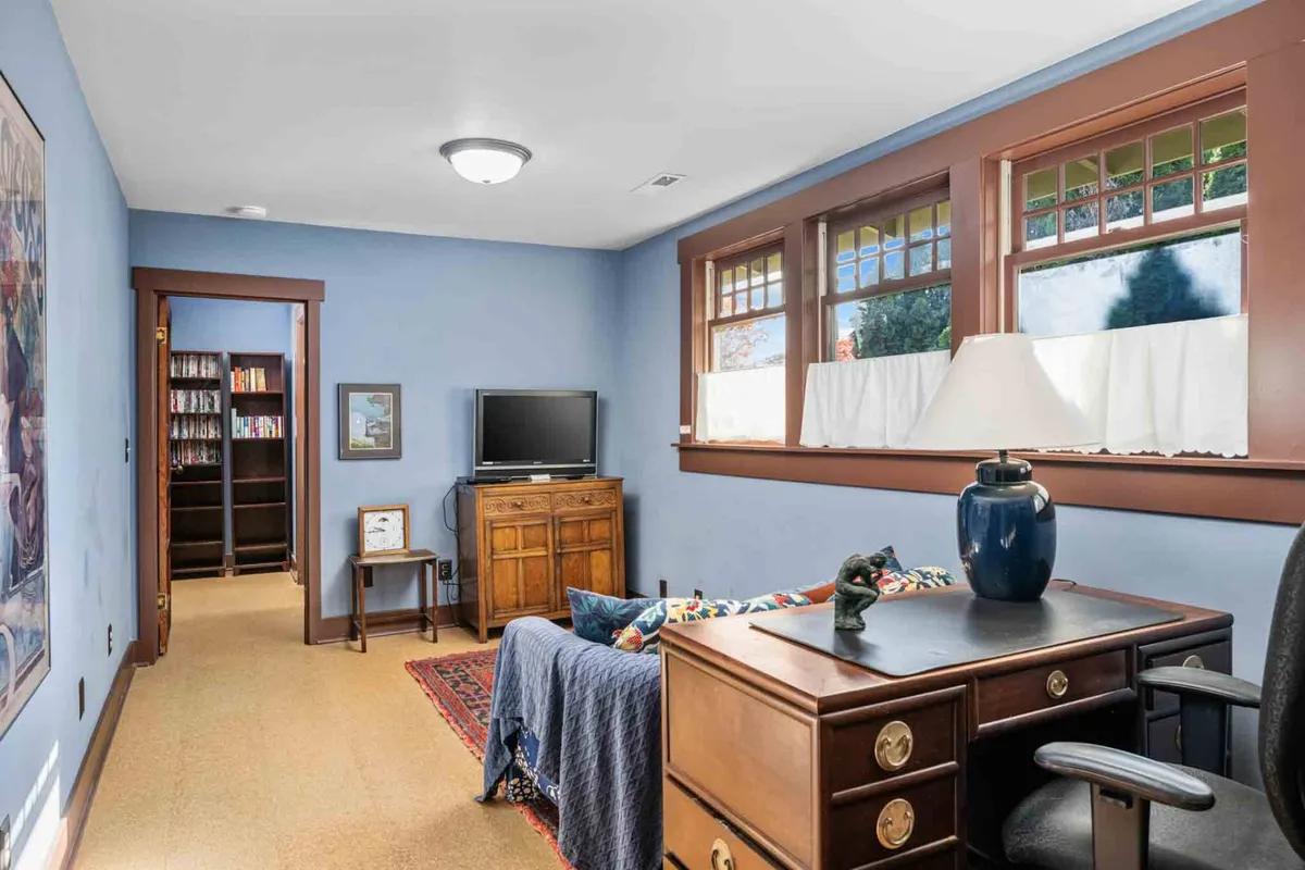 A serene office den lit by soft daylight from a curtained window, featuring a wooden desk with a lamp, a wall-mounted bookshelf filled with books, and framed artwork. The space is designed for quiet reflection or productive work.