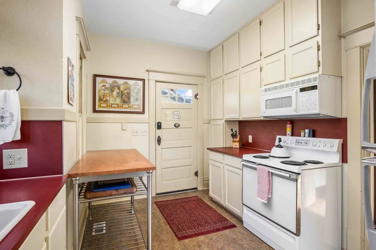 A charming kitchen featuring a classic white stove, vibrant red countertop, and a welcoming wooden table set near a door with a window that hints at easy access to the outdoors. The space feels homey and functional, ideal for preparing meals or blending work and relaxation.
