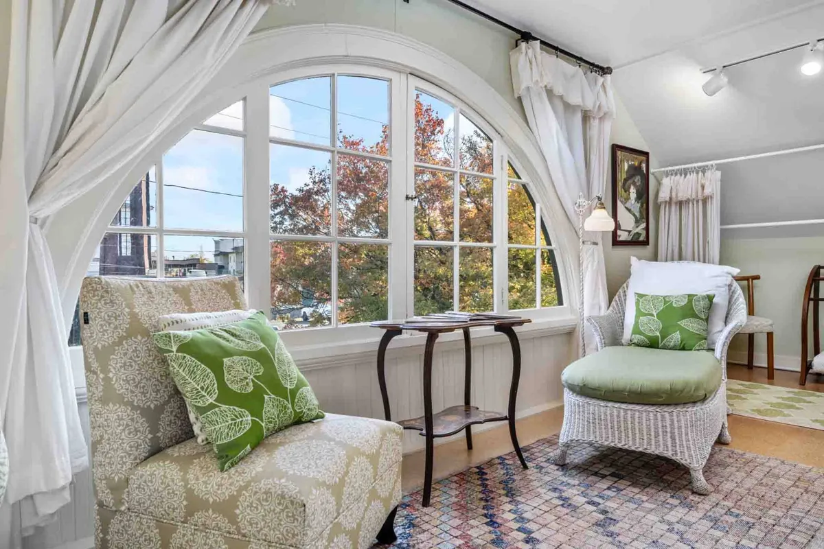 Inside the home, a spacious nook is lit by a large arched window. Wicker chairs, green accent pillows, and a small side table create a calm, sunlit spot for reading, sipping coffee, or chatting. The space is bright and welcoming, ideal for candid group photos and relaxed gatherings.