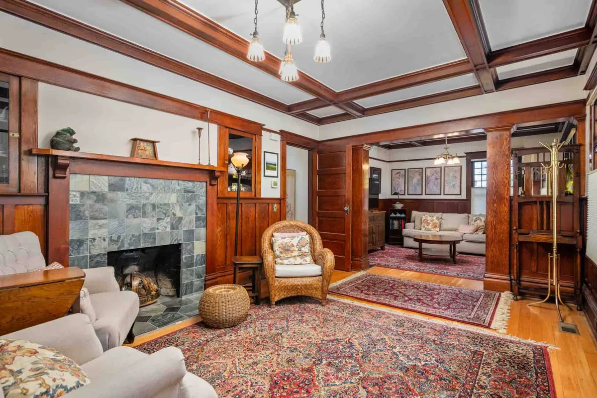 A warm, inviting living room with rich wood paneling, a glowing fireplace, plush armchair, and a stylish rug create a cozy space ideal for relaxing or socializing. Artistic displays adorn the walls, adding a touch of historic elegance to the scene.