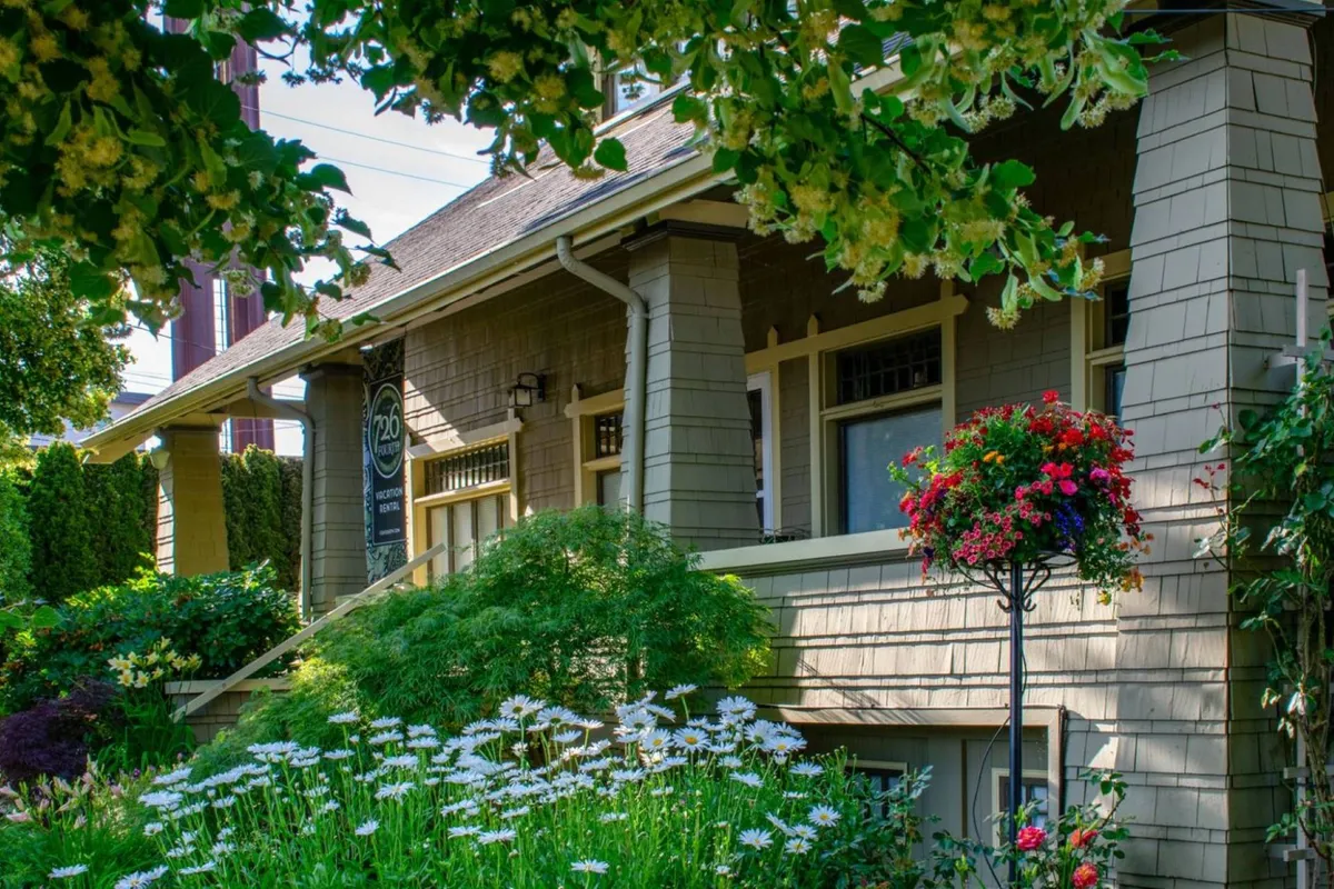 This image shows the front porch of the historic home, bordered by lush flower beds and a charming hanging flower basket. The porch features comfortable seating and a welcoming atmosphere, perfect for morning coffee or gathering with friends. The overall vibe is inviting and picturesque, ideal for socializing and relaxing outdoors.