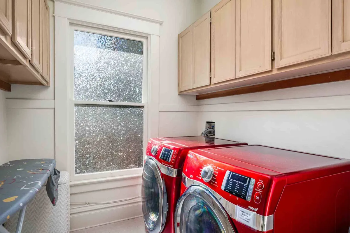 A practical laundry room featuring a red washing machine and dryer beneath a window with textured glass. The space is organized and well-lit, ideal for tackling laundry tasks during an extended stay.